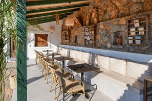 Een terras met rieten stoelen en tafels, een stenen muur met boekenplanken en een houten deur.