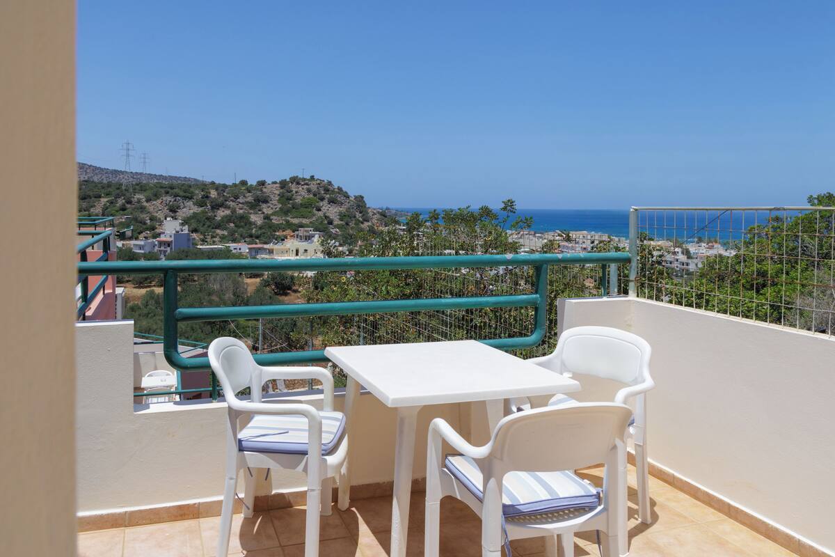 Un balcon avec une table et des chaises blanches, une vue sur la mer et une rambarde verte.