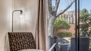 Una stanza con una sedia decorata, un letto e vista su un cortile alberato con un edificio.