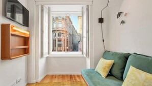 A cozy room with a green sofa, a wooden floor, a window with a view of buildings, and a mounted TV.