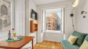 A room with a wooden table, a green sofa, a window with a view of buildings, and a mounted TV.