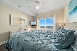 Quarto com cama, ventoinha no teto, televisão, janela com vista e um quadro na parede.
