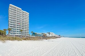 Un hôtel situé en bord de mer, doté de nombreux balcons, d’une plage de sable et offrant une vue sur un ciel bleu azur.