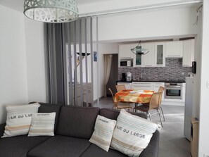 A modern living room with a sofa, a dining table with chairs, and a kitchen in the background. - House with a fenced-in yard, a 5-minute walk from the beach, next to the forest   (La Barre-de-Monts)