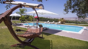 A hammock chair by a pool with lounge chairs and umbrellas, surrounded by greenery and a view of distant mountains. - Villa Alessia 6 in Gela (Gela)