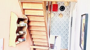 Un pasillo estrecho con una escalera de madera que lleva a una plataforma. Hay estanterías a la izquierda y un cuadro enmarcado a la derecha.