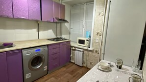 A compact kitchen with purple cabinets, a washing machine, a microwave, and a dining table set with plates and cutlery. - Daniko (Taraz)