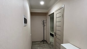 A narrow hallway with a light-colored floor, a coat rack, a door with a mirror, and a white dresser. - Daniko (Taraz)