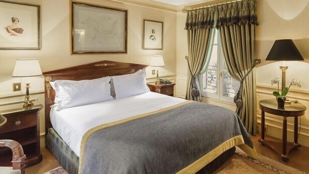 A neatly arranged hotel room with a large bed, two bedside tables with lamps, a window with curtains, and framed artwork on the walls. - Daniko (Taraz)