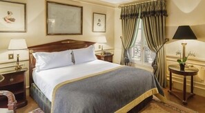 A neatly arranged hotel room with a large bed, two bedside tables with lamps, a window with curtains, and framed artwork on the walls. - Daniko (Taraz)