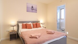 Un letto rifatto con cura, una coperta e cuscini rosa, un comodino con una lampada e un quadro raffigurante un faro sulla parete.