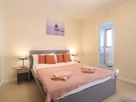 Una cama bien hecha con una manta y almohadas rosas, una mesita de noche con una lámpara, y un cuadro de un faro en la pared.