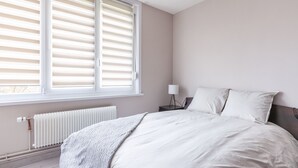 A bedroom with a large window, a radiator, a bedside table with a lamp, and a bed with white bedding.