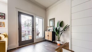Une pièce spacieuse dotée d’une porte-fenêtre coulissante donnant sur un balcon, d’un plancher en bois, d’un mur blanc orné d’un miroir à carreaux et d’une plante en pot.