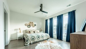 Une chambre à coucher comprenant un lit, un ventilateur de plafond, une table de chevet, une lampe, un mur décoré d’assiettes et de grandes fenêtres avec des rideaux.