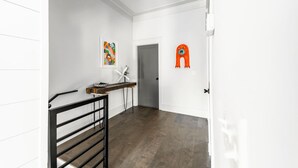 Un couloir avec une porte blanche, une rambarde en métal noir, une petite table sur laquelle se trouve un bol, et une peinture abstraite aux couleurs vives accrochée au mur.