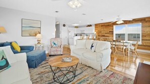 Ein modernes Wohnzimmer mit einem blauen Sofa, einem weißen Sofa und einem Couchtisch. Es gibt einen Essbereich mit einem Holztisch und Stühlen. Im Hintergrund ist eine Küche mit weißen Schränken und einer Mikrowelle erkennbar.