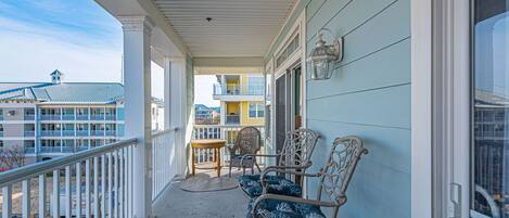 A covered balcony with wicker chairs and a table, overlooking a residential area with multiple buildings.