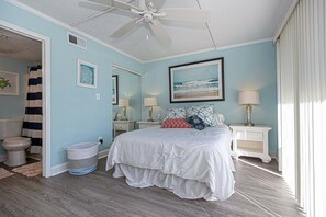 A bedroom with a bed, bedside tables, a ceiling fan, a large window, and a bathroom visible through an open door. - 2 bedroom Ocean Block condo, with 2 parking spaces and free Wifi, located in midtown and just a few steps from the beach! (Ocean City)