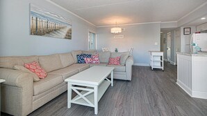 Sala de estar com sofá seccional bege, mesa de centro branca e pintura de uma cena de praia. Há uma área de cozinha com geladeira e pia.