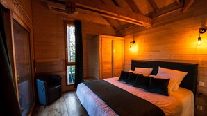 A wooden cabin bedroom with a bed, a small chair, a window, and a wall-mounted light.