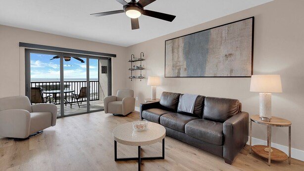 Un salon moderne comprenant un canapé, deux fauteuils, une table basse et un ventilateur de plafond. Une grande fenêtre offre une vue sur l’extérieur.