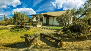 Un edificio rustico con tetto in tegole, circondato da un vialetto sterrato, alberi e una piccola costruzione in legno.