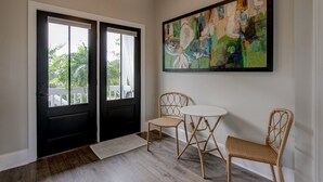 Une pièce avec une porte noire, un mur blanc et un grand tableau abstrait. On y trouve deux fauteuils en osier et une petite table ronde.