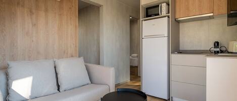 Un espacio habitable compacto con sofá, una pequeña mesa redonda, una kitchenette con microondas y refrigerador, y un muro decorativo de madera.