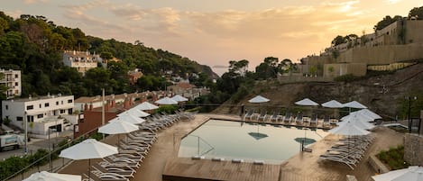 A pool area with lounge chairs and umbrellas, surrounded by buildings and a hilly landscape.