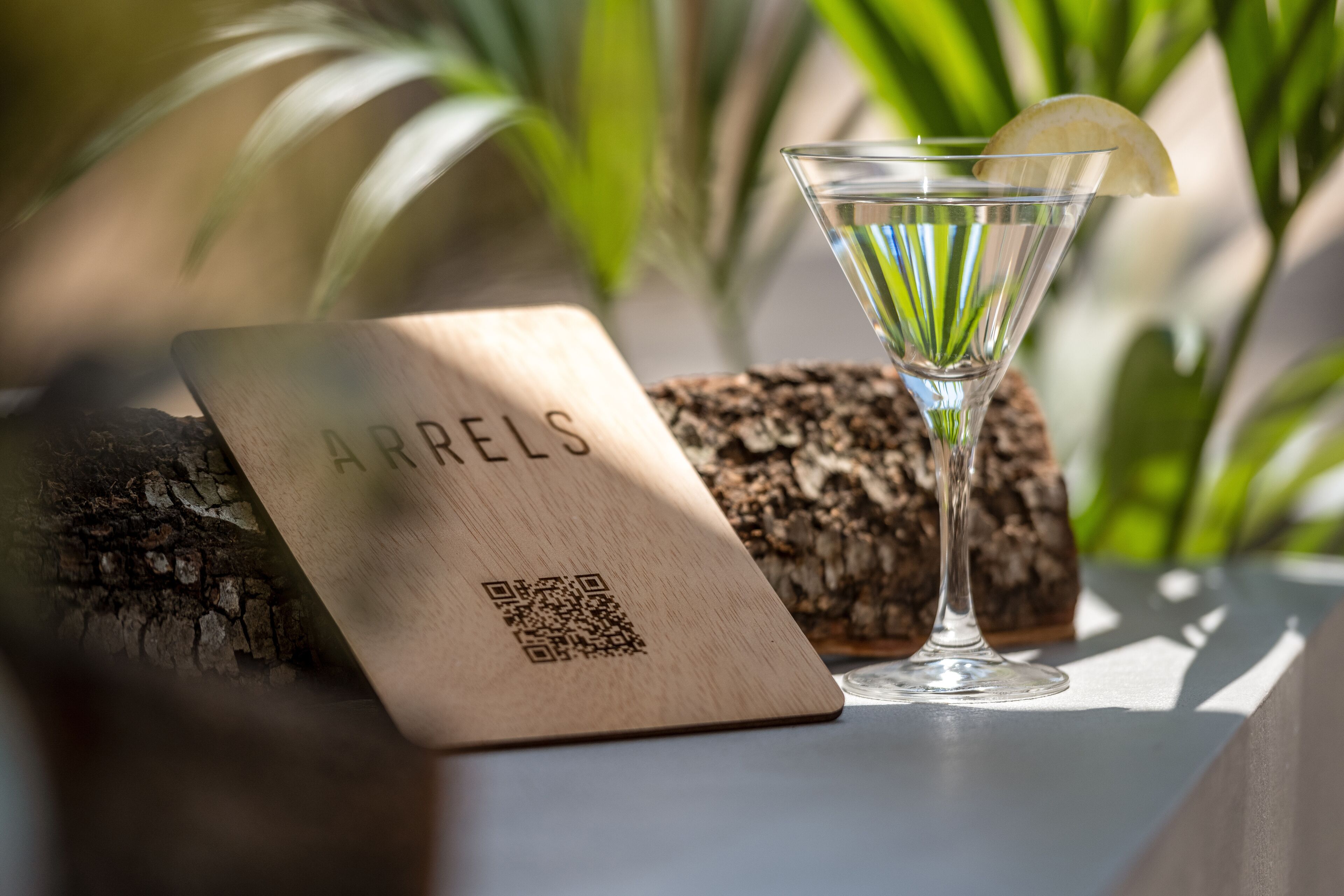 Una placa de madera con un código QR y la palabra "ARRELS" sobre una mesa junto a una copa de martini con una rodaja de limón.