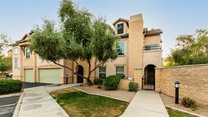 A two-story residential building with a beige facade, a green garage door, and a landscaped front yard with a tree and shrubs. - Surprise Ground Floor Condo with 2 Private Courtyards! Heated Community Pool! (Surprise)
