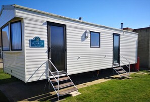 Uma casa móvel com uma placa indicando 'Sunrise 28 x 10 MxIII', uma porta da frente, duas janelas e um conjunto de escadas externas.