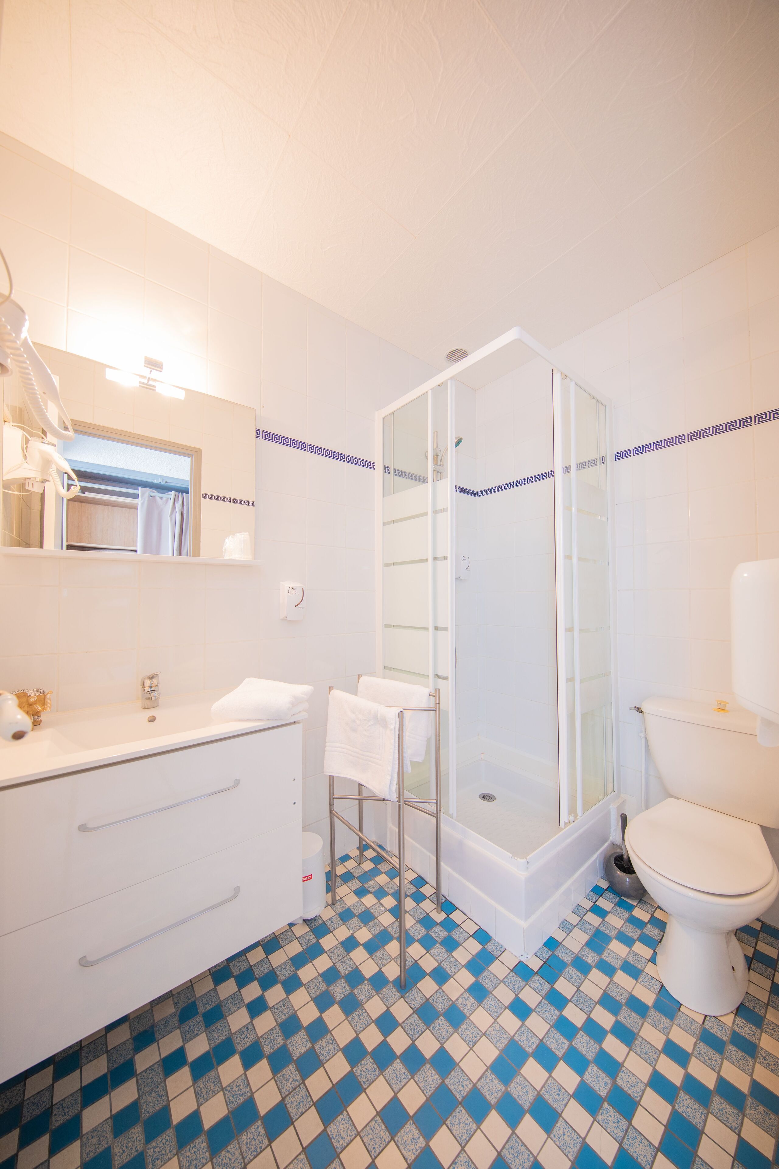 Une salle de bain avec un lavabo blanc, un miroir et des toilettes, une douche en verre et un carrelage à motifs à carreaux.