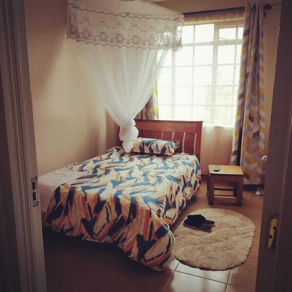 A bedroom with a bed, a nightstand, a chair, and a window with curtains. - Stay.Plus Shaba Village Apartment (Syokimau)
