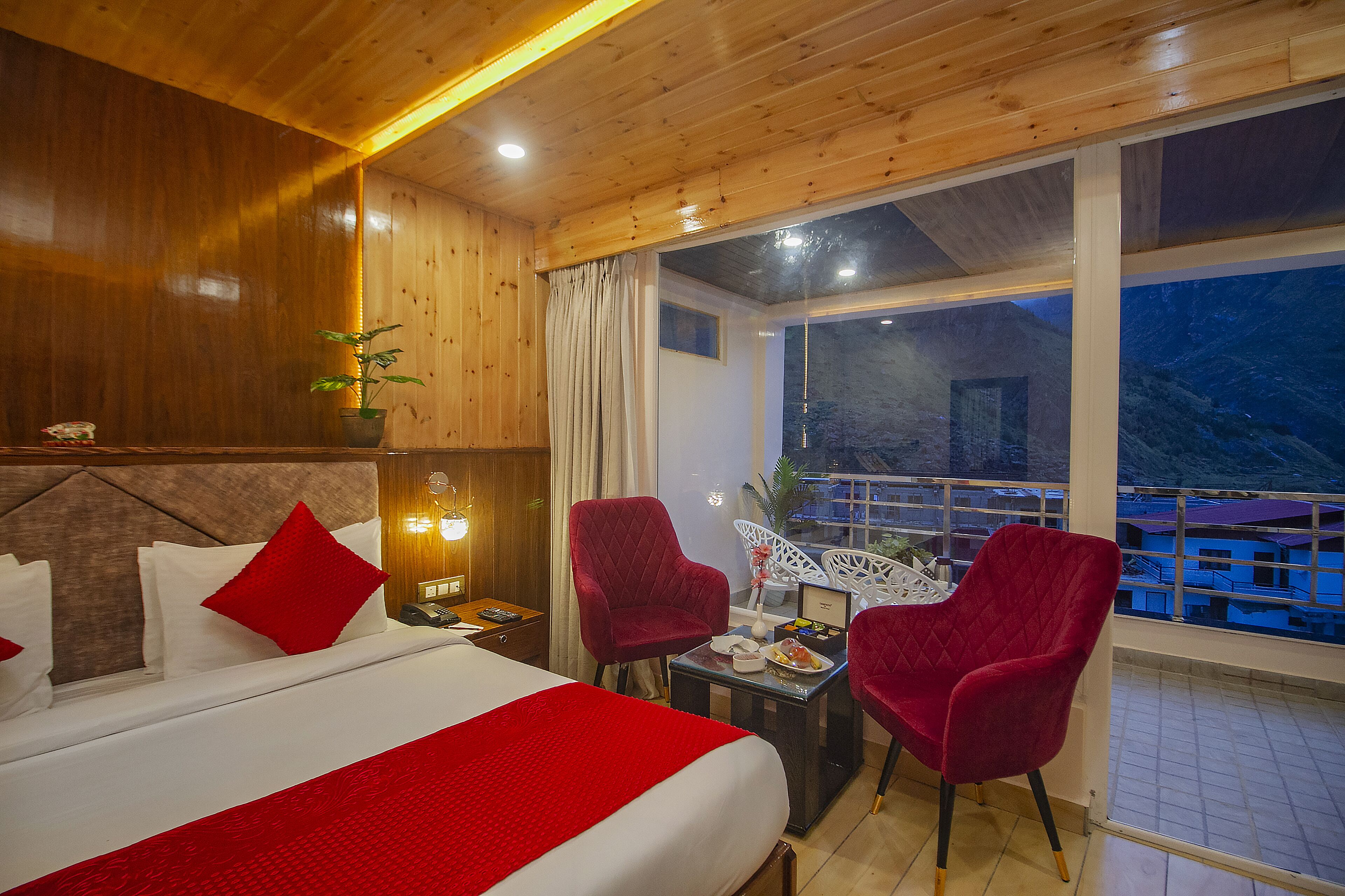 A hotel room with a wooden ceiling, a large bed with red bedding, two red chairs, a small table with a lamp, and a view of a balcony with buildings.
