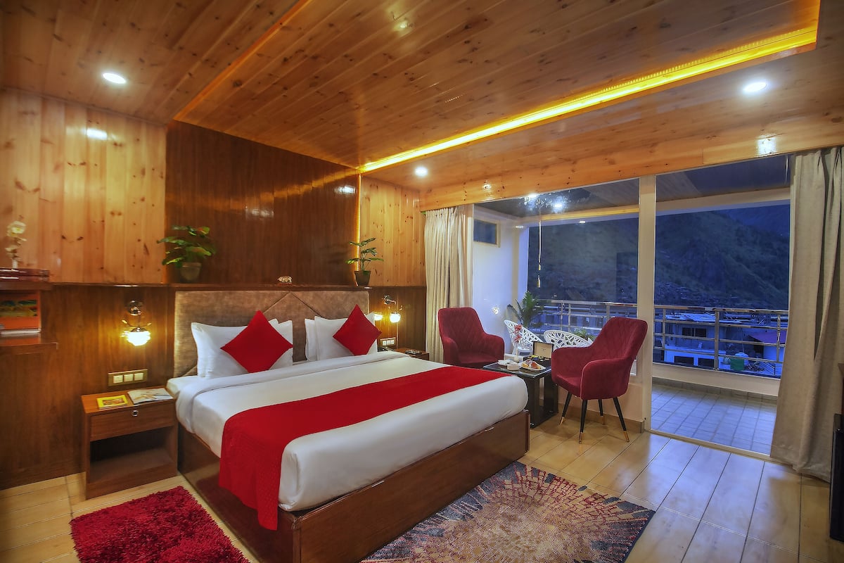 A wooden-paneled bedroom with a large bed, red accents, a balcony with a view, and a nightstand with a lamp.