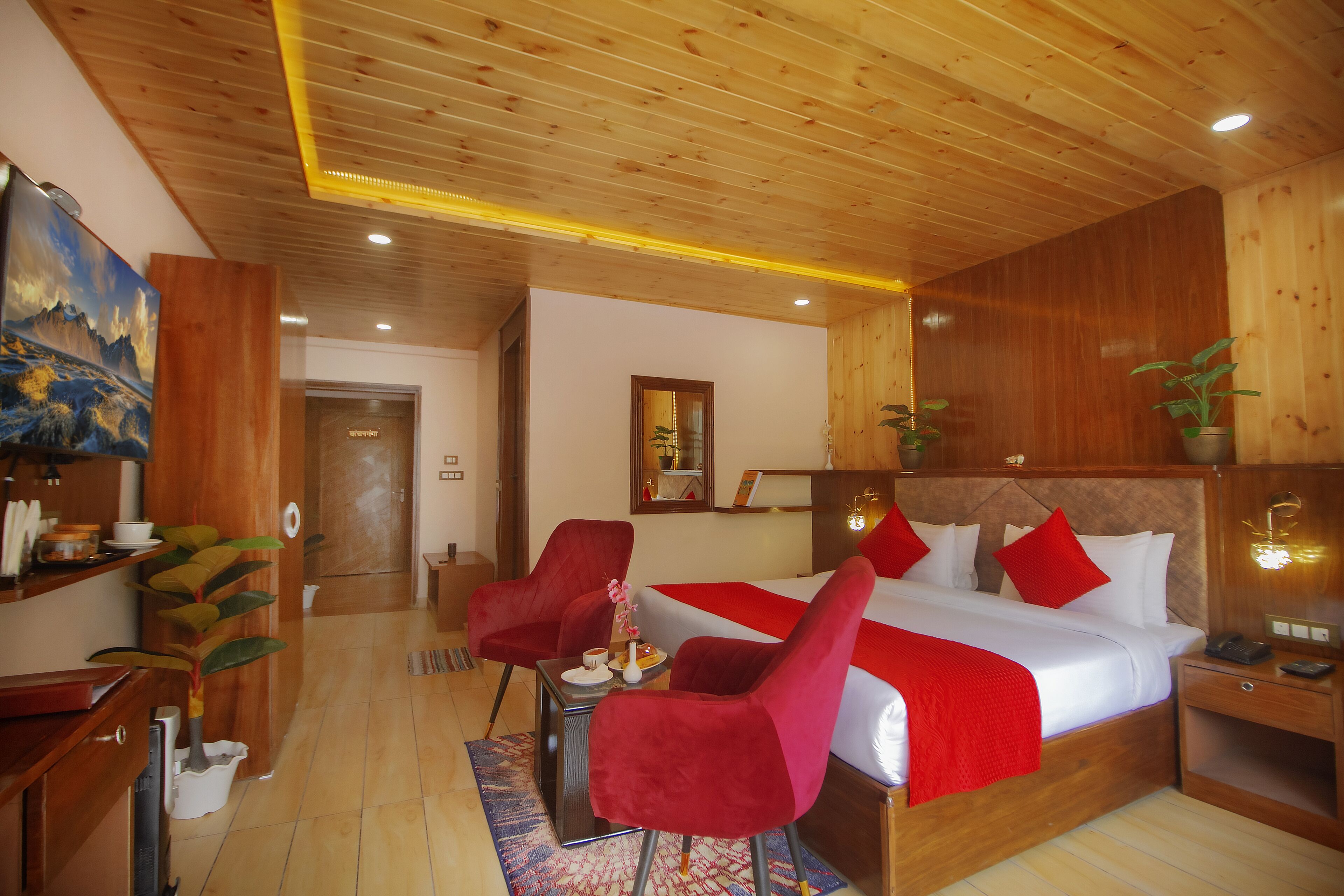 A hotel room with a wooden ceiling, a large bed with red bedding, two red chairs, a small table with a vase, and a wooden nightstand.