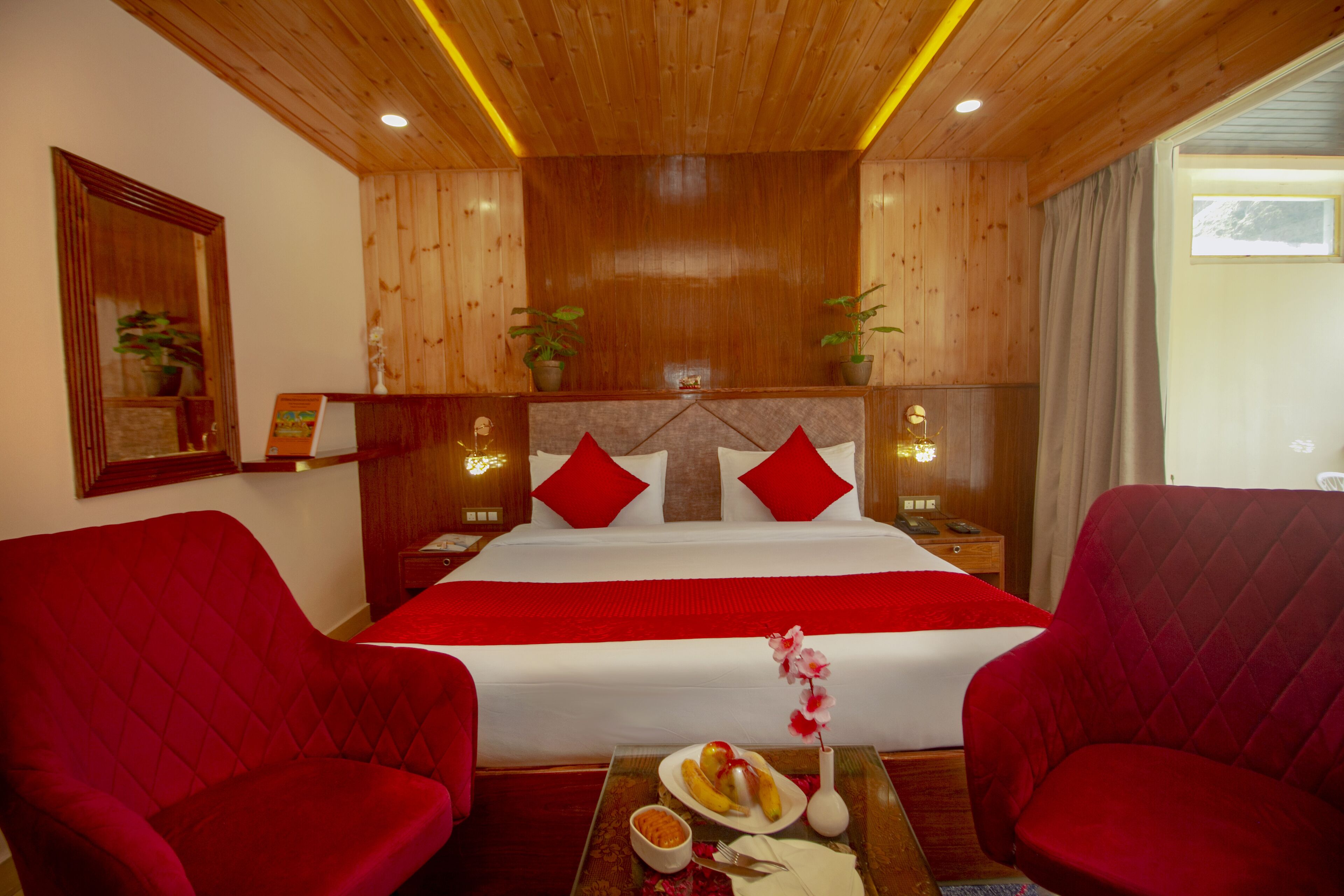 A hotel room with a large bed, two red upholstered chairs, a small table with a plate of fruit and a vase of flowers, and wooden paneling on the walls.