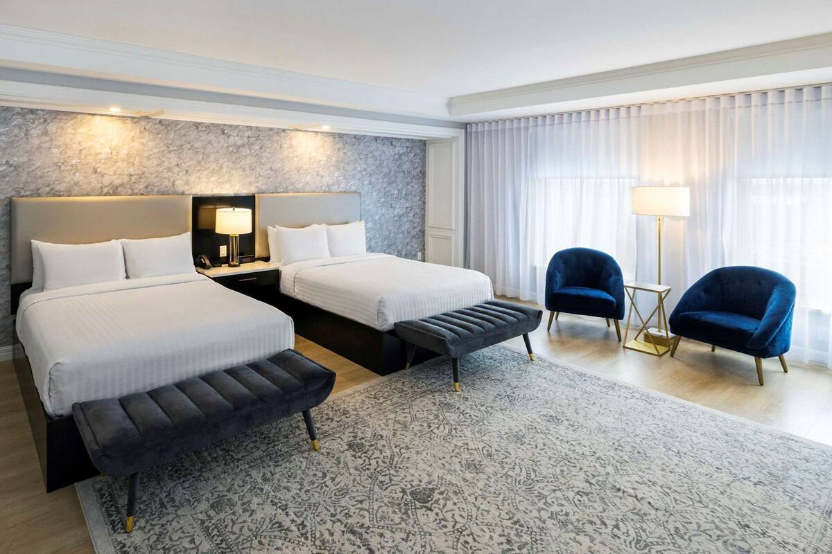 A hotel room with two beds, a bench, two armchairs, a lamp, and a large window with curtains.