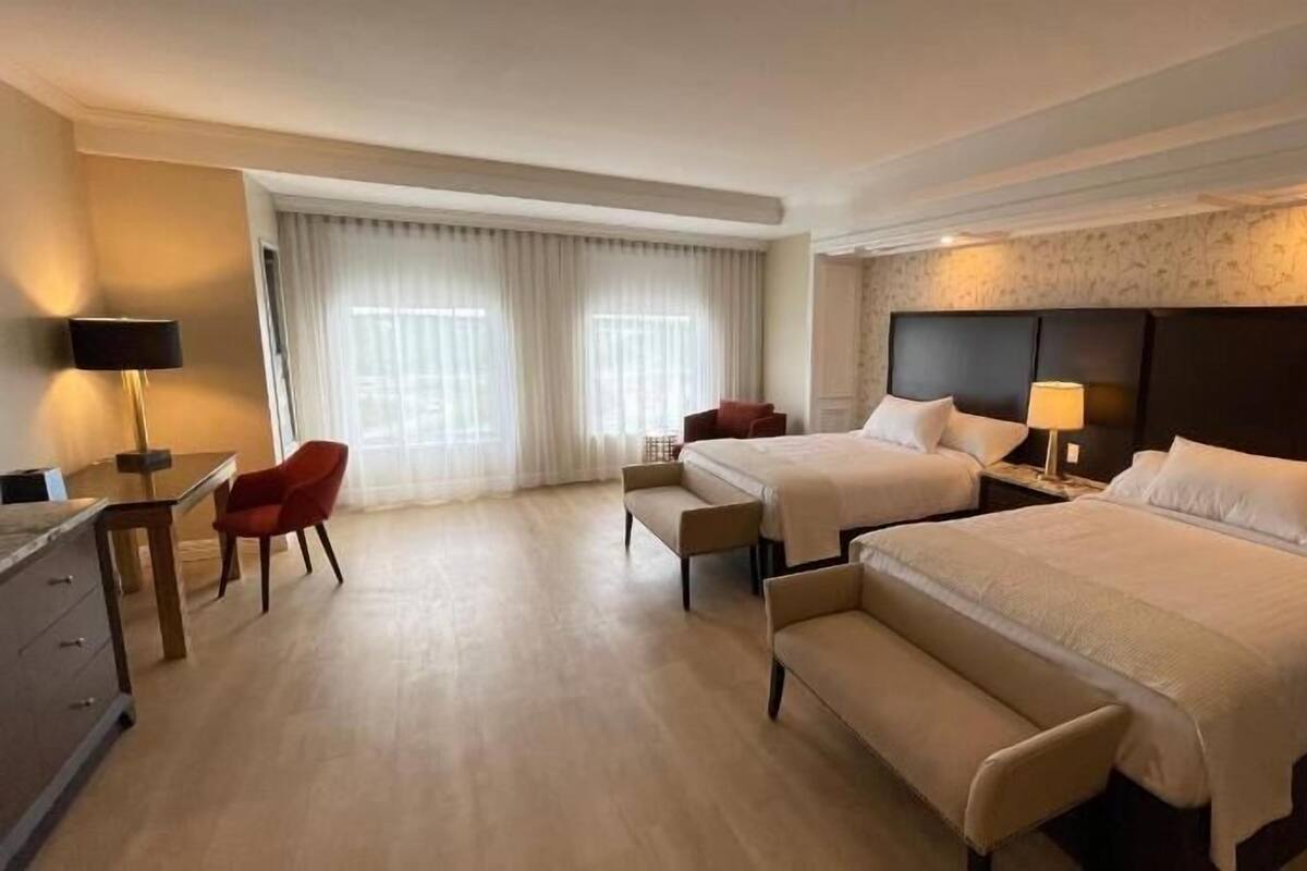 A hotel room with two beds, a desk, a chair, and a large window with curtains.