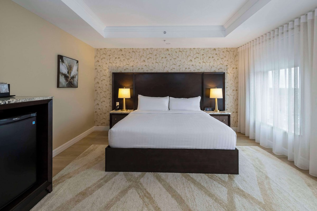 A hotel room with a large bed, two bedside lamps, a nightstand, a desk with a computer, and a large window with sheer curtains.