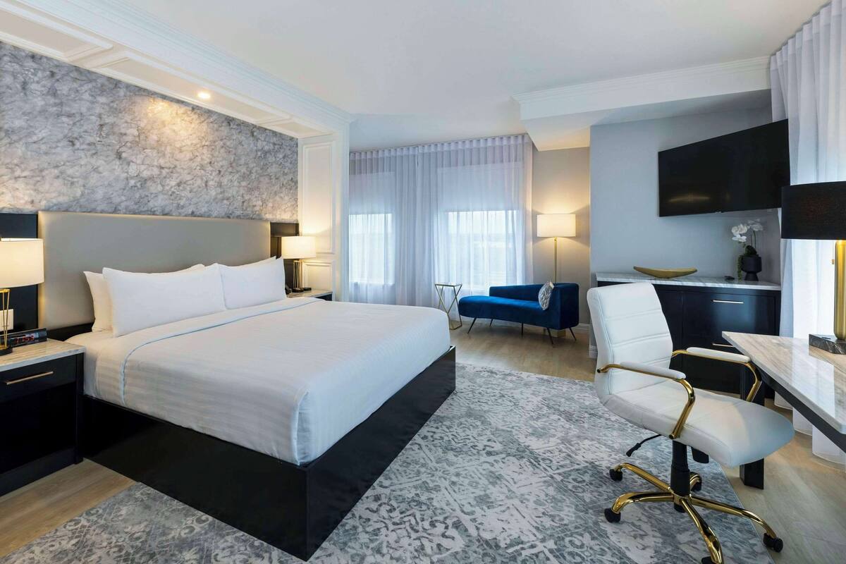 A modern hotel room with a large bed, a desk with a chair, a flat-screen TV, and a large window with curtains.