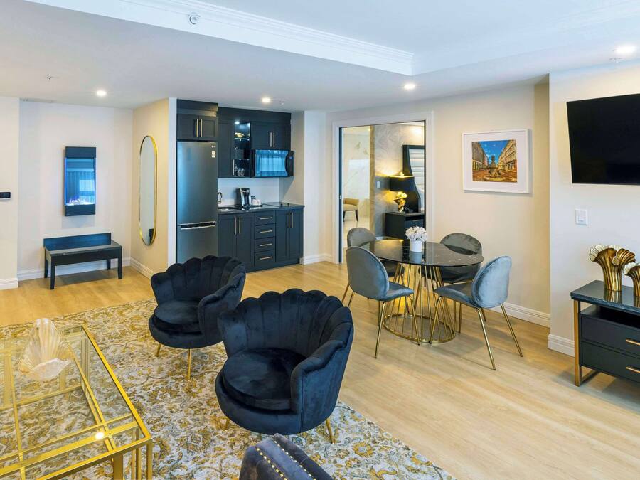 Een moderne woonkamer met een glazen salontafel, twee zwarte fauteuils, een eethoek met een ronde tafel en stoelen, een keuken met donkere kastjes, en een flatscreen televisie aan de muur.