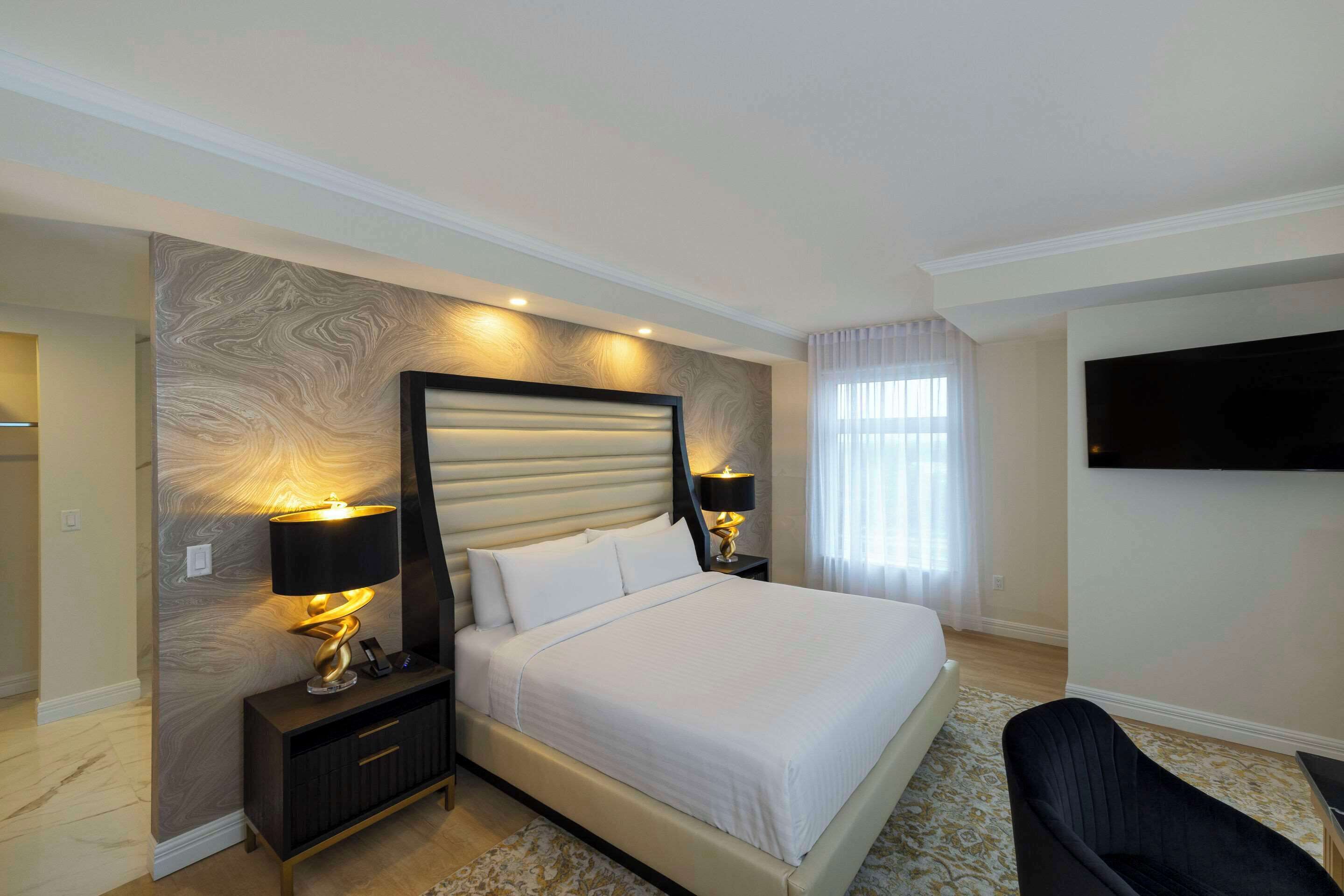A hotel room with a large bed, two bedside lamps, a flat-screen TV mounted on the wall, and a dark-colored armchair.