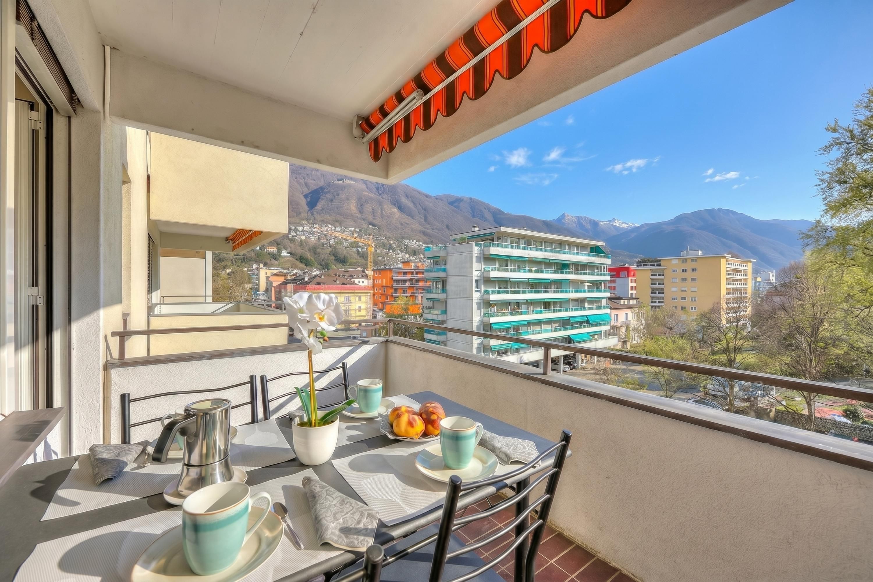 Un balcón con una mesa dispuesta para dos, una planta en maceta y vistas a edificios y montañas.