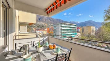 A balcony with a table set for two, a potted plant, and a view of buildings and mountains.