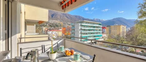 Un balcón con una mesa dispuesta para dos, una planta en maceta y vistas a edificios y montañas.