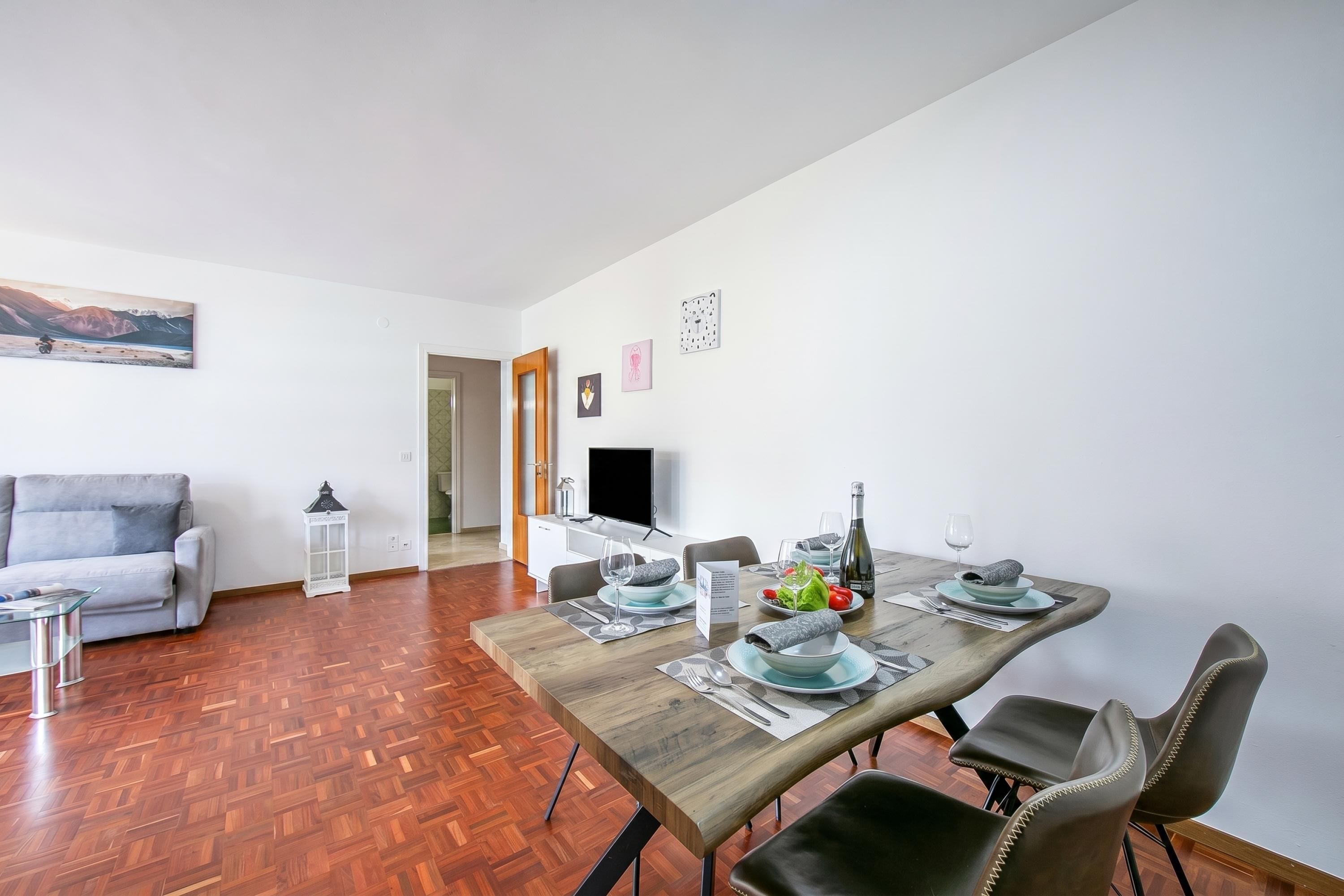 A dining area with a wooden table set for four, a grey sofa, and a TV in the background.