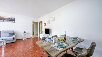 A dining area with a wooden table set for four, a grey sofa, and a TV in the background.